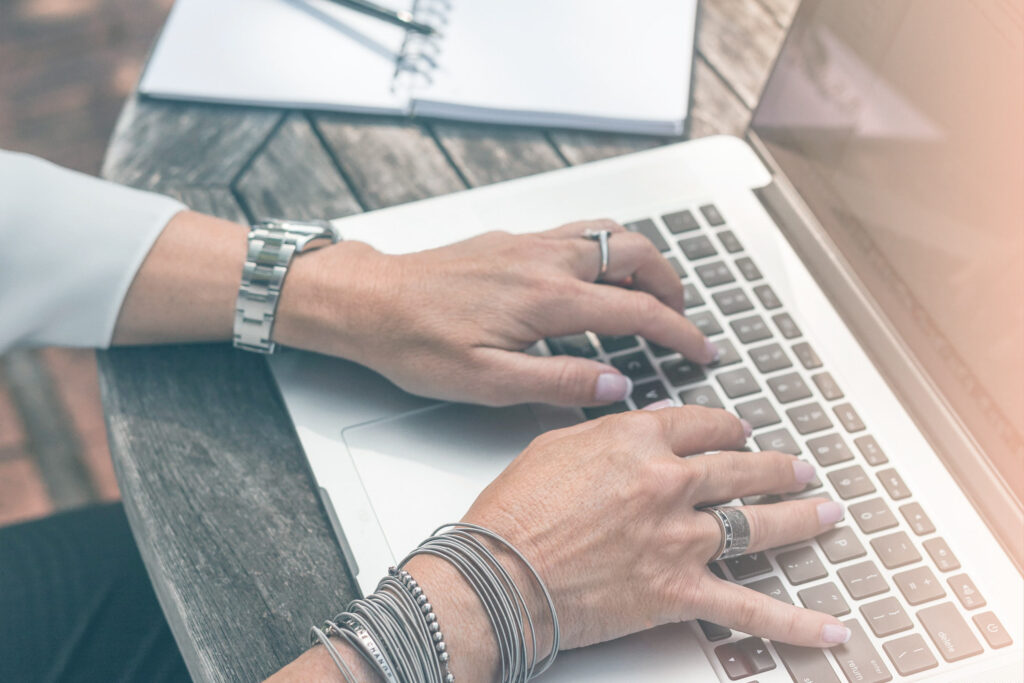 Deux mains de femme avec des bracelets qui tapent sur un ordinateur portable, un cahier et crayon sont posés sur le bureau.