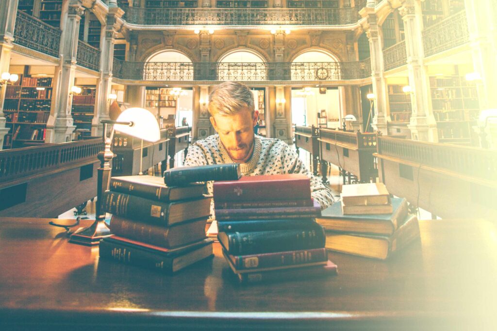 Homme blond concentré dans une bibliothèque monument avec une pile de livre devant lui et une lampe de table.