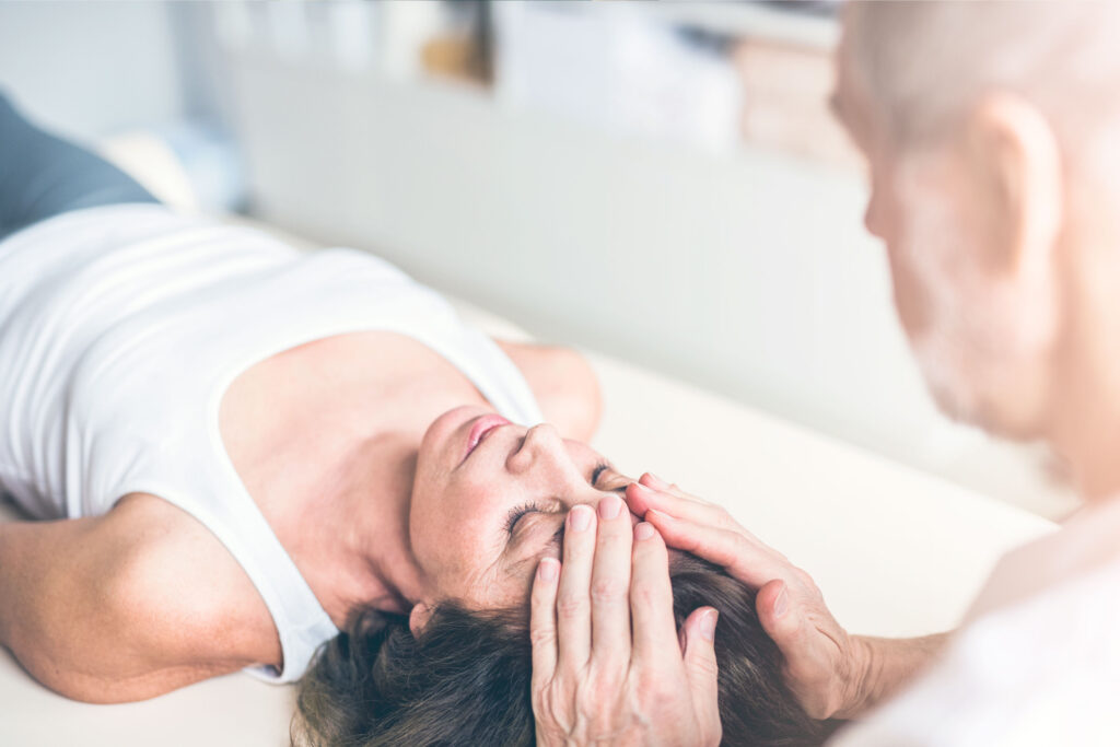 Homme mature aux cheveux gris qui fait un massage de tête à une femme aux cheveux bruns, couchée sur un lit de massage.